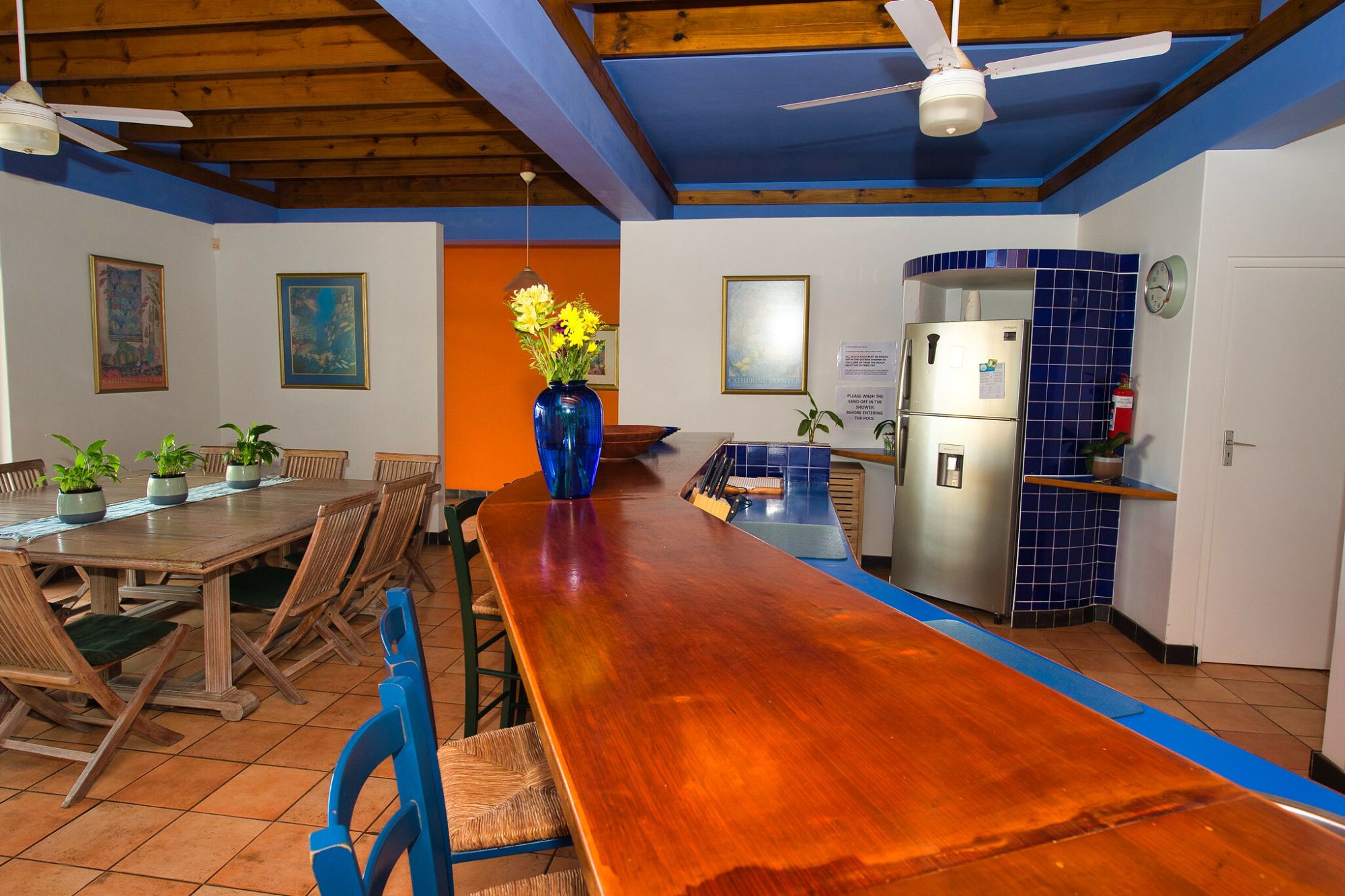 A bar with wooden counter top. The bar chairs are wooden, with white back rests. There is a blue vase with yellow flowers. A silver fridge stands to the back right of the image. There are 2 white ceiling fans. The ceiling is blue and wooden.